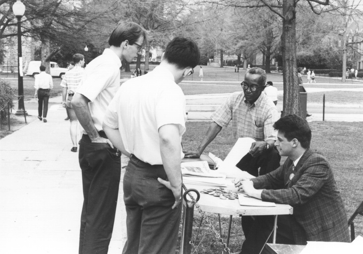Two white men stand at an information table. A white male student and Oliver Harvey are behind the table. 