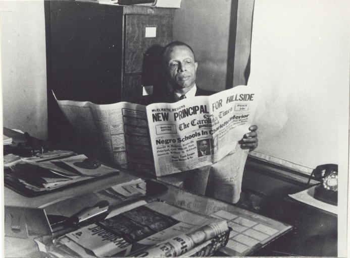 Louis Austin, seated behind a desk, reads an issue of the Carolina Times.