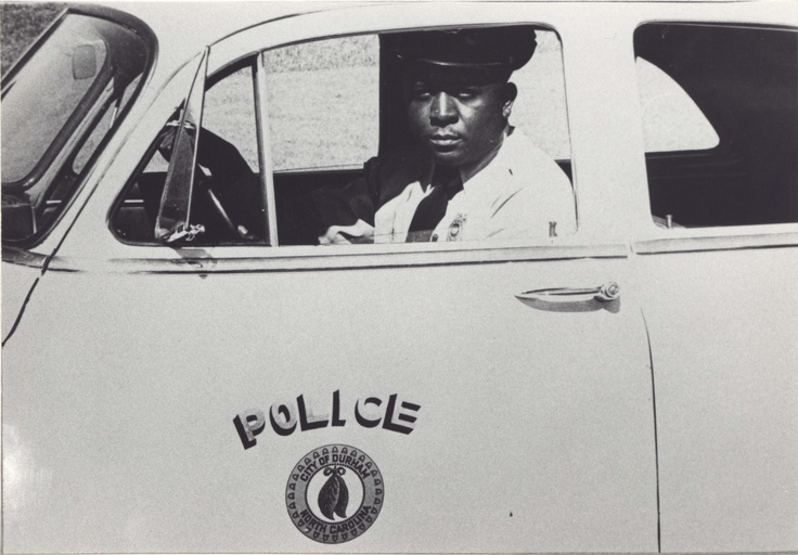 A black police officer is seated in the driver's seat of a police car. 