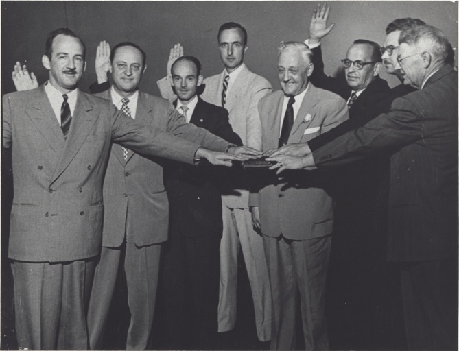 A group of men hold their hands on a book and raise their other hands in the motion of taking an oath. 