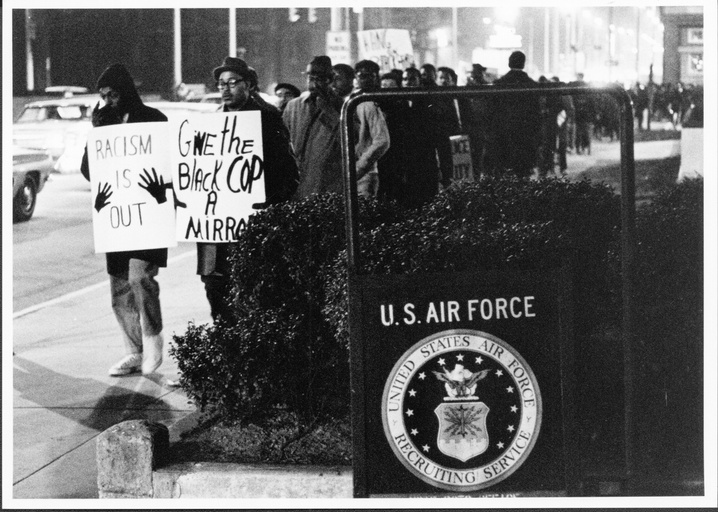 A long line of protesters holding signs. A sign for the US Air Force Recruiting Station is visible. 