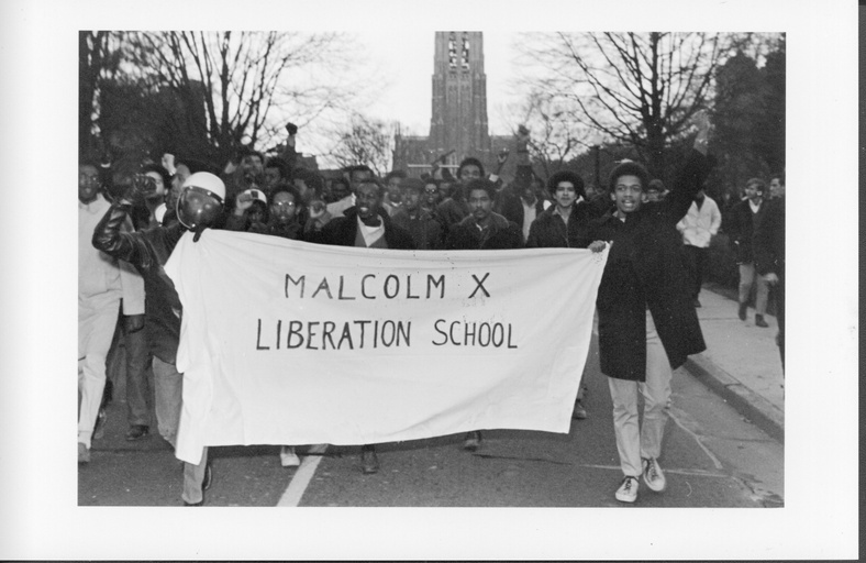 A large group of primarily African-American people with the Duke Chapel in the background. They carry a banner that reads "Malcolm X Liberation School."
