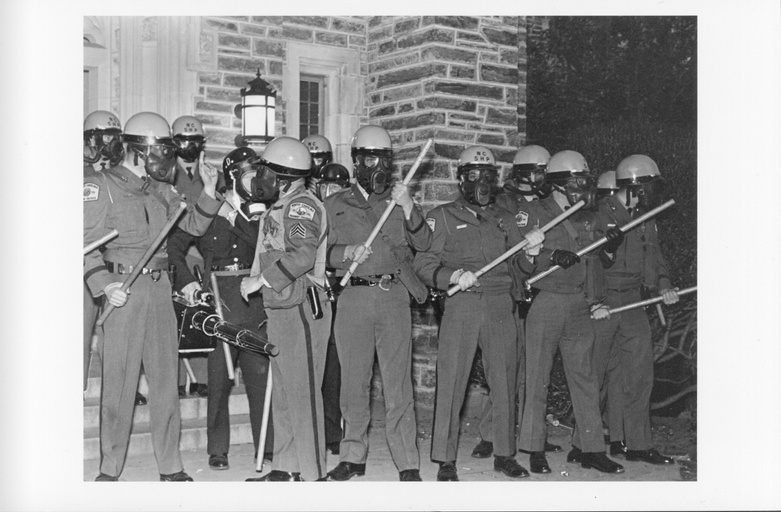 A group of officers wearing gas masks and carrying batons stand together. 