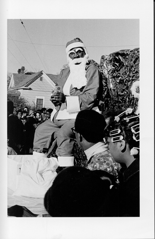 An African-American man dressed as Santa Claus sits atop a float. Numerous people are gathered around him. 