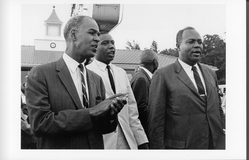 From left to right: Roy Wilkins, Floyd McKissick, and James Farmer in suits standing outside. A fourth man is standing behind them with his back turned. 