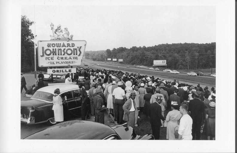 A large group of people gather along the side of a highway outside a Howard Johnson's.