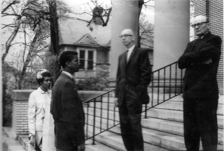 A black man and Fay Mayo stand on the stairs outside a church facing two white men. 