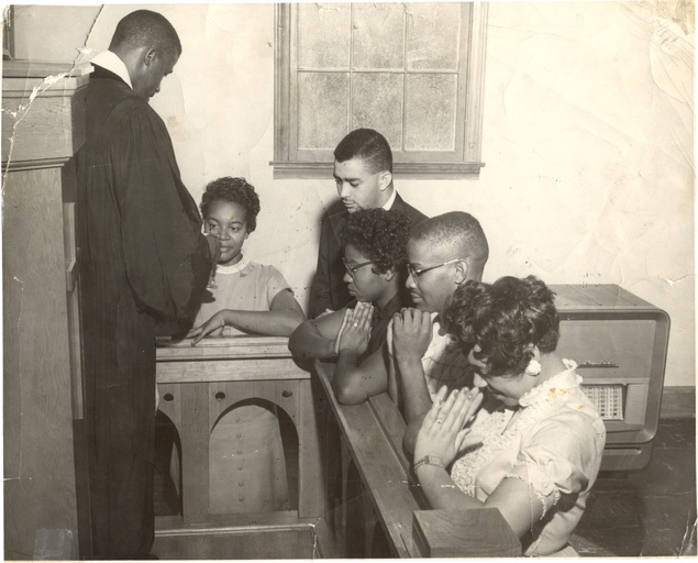 Five black men and women kneel in prayer before a black minister.