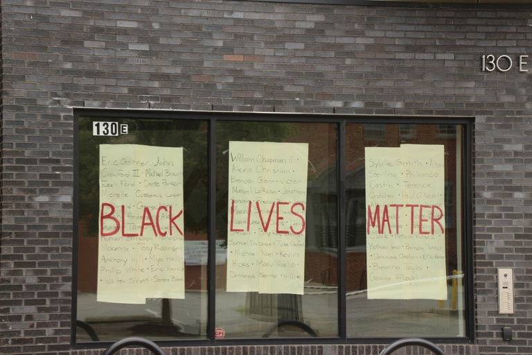 Three window panels with yellow paper hanging in them. Names of black people murdered by police are written on the paper. The words "Black Lives Matter" are painted in red across the windows.
