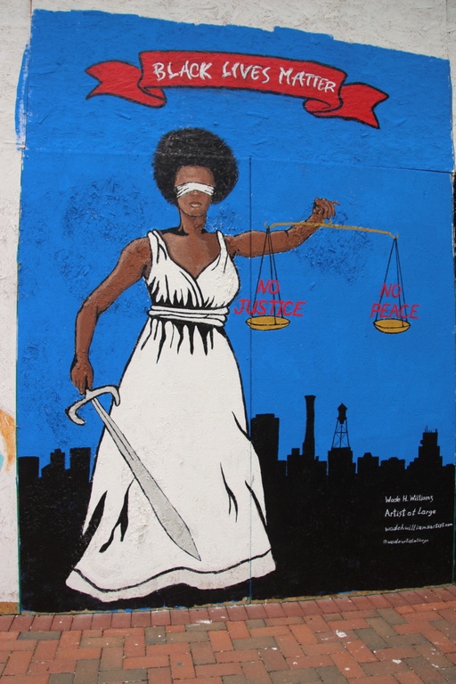 A mural with a black woman depicted as lady justice holding scales with the words "no justice no peace" and a red banner above her head displaying the words "black lives matter."